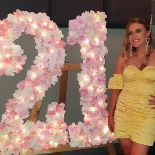 A 21st birthday girl standing next to a sparkling '21' sign, surrounded by loved ones, celebrating this significant milestone.